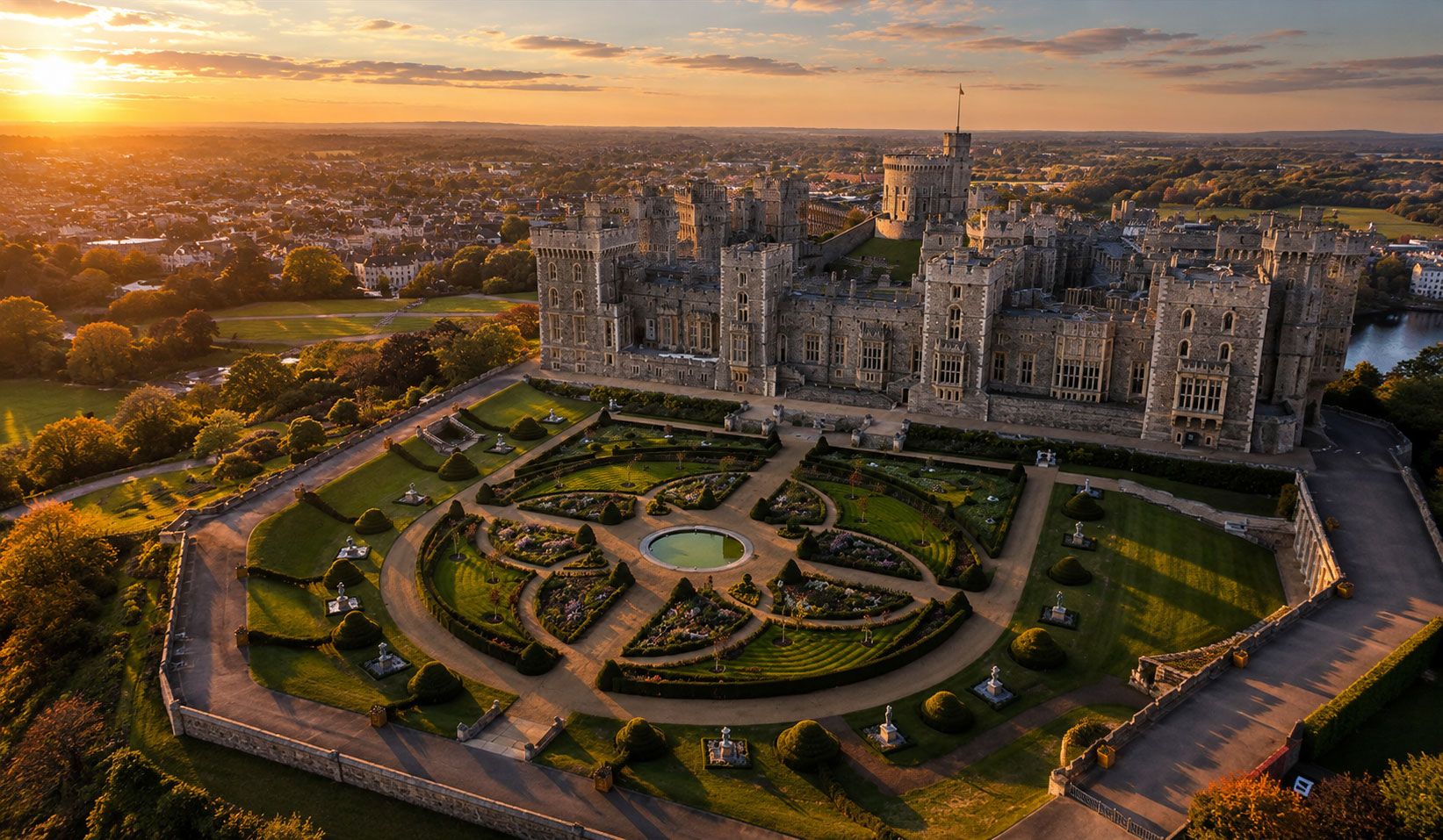 Aerial view of a grand medieval stone castle and its formal gardens at golden hour, photographed from an angled perspective.